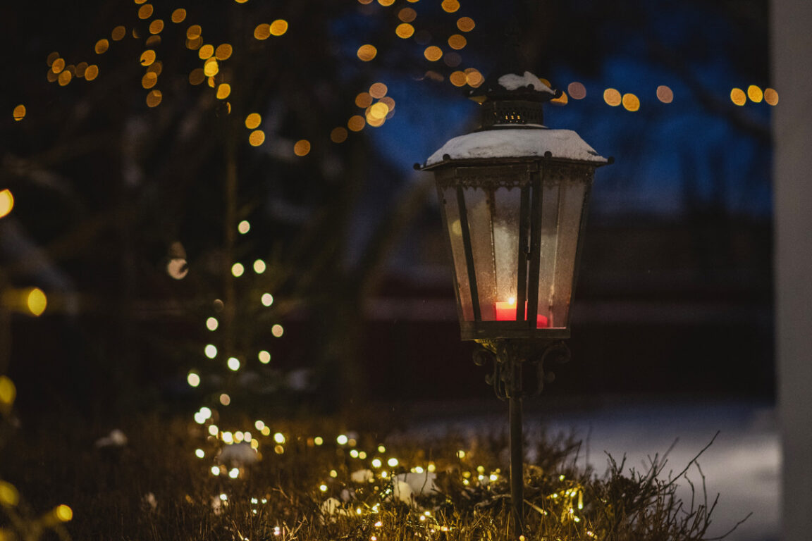 En lykta med ett rött ljus i fotograferat i skymningen. I bakgrunden syns julljus.